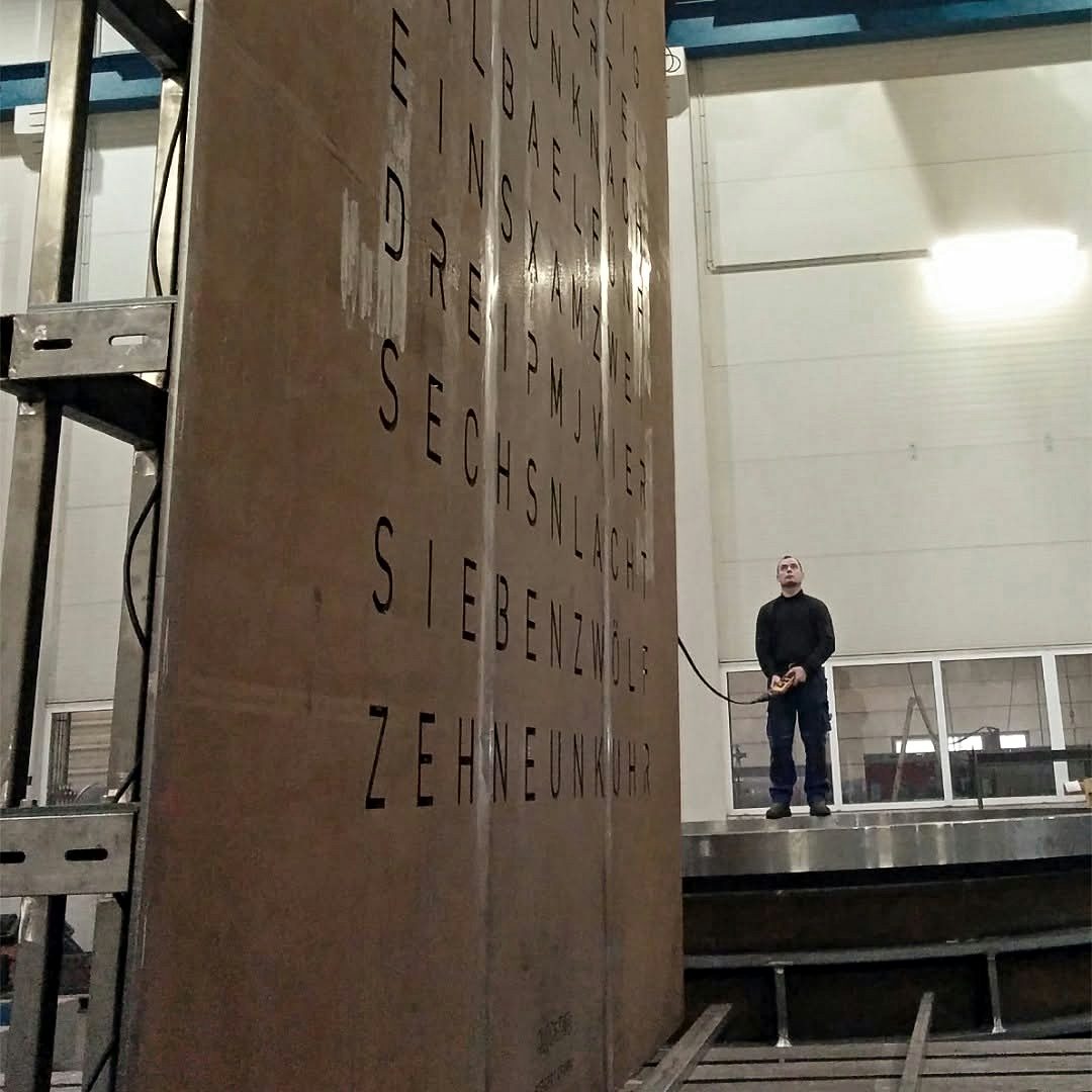 Aufgerichtete Cortenstahl-Frontplatte der größten Wortuhr der Welt in der Fertigung von Bock Maschinenbau, eine Person daneben als Größenvergleich