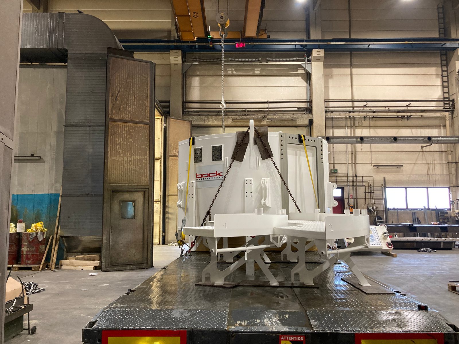 Coated cylindrical machine housing and curved platform segments with tubular handrail, loaded onto the low loader by overhead crane